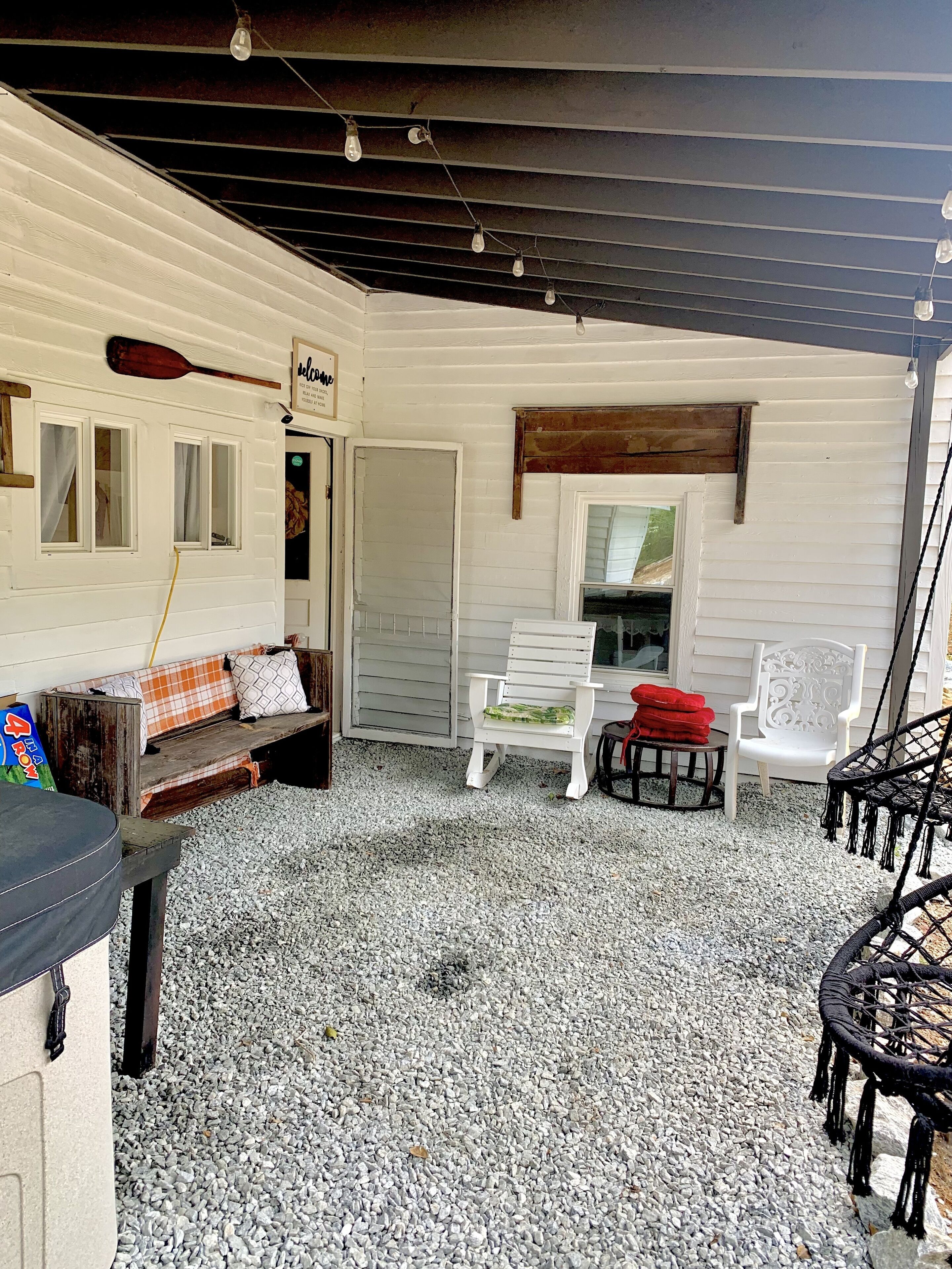 A covered patio area with a white bench, a white chair, and a wicker chair. There is a wooden paddle on the wall and a small table with a red cloth.