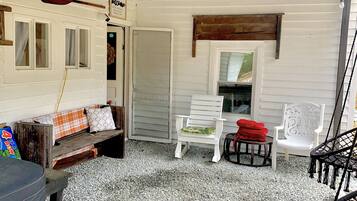 A covered patio area with a white bench, a white chair, and a wicker chair. There is a wooden paddle on the wall and a small table with a red cloth.