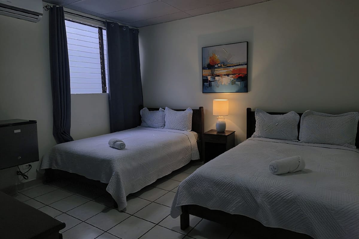Ein Hotelzimmer mit zwei Betten, einem Schreibtisch, einem Stuhl, einer Lampe und einem Fenster mit Vorhängen.