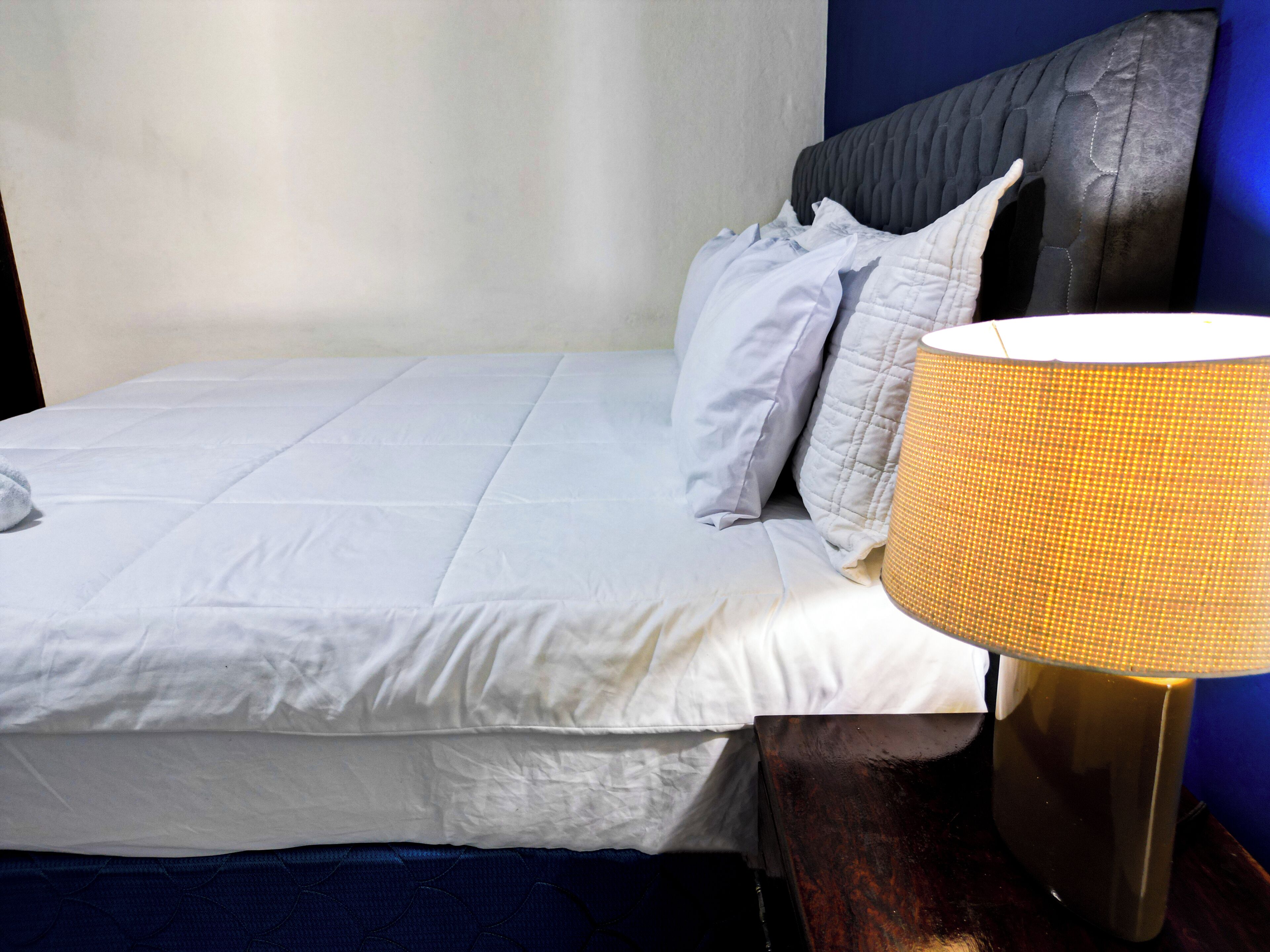 A neatly made bed with white bedding and a dark headboard, a lamp with a golden base, and a blue wall in the background.