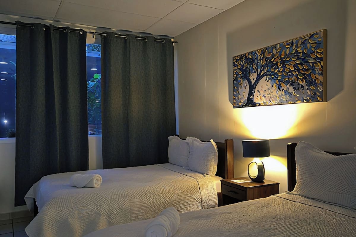 Ein Hotelzimmer mit zwei Betten, einem Nachttisch mit Lampe, einem Wandbild und Blick ins Freie durch das Fenster.