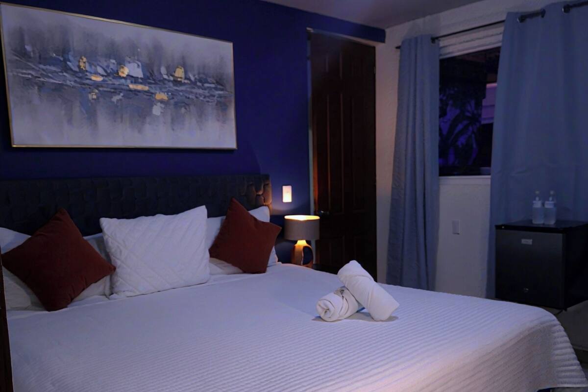 Ein Hotelzimmer mit einem großen Bett, einem Gemälde an der Wand, einem Nachttisch mit einer Lampe und einem Fenster mit Vorhängen.