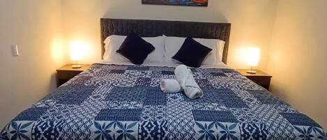 A neatly made bed with a patterned bedspread, two pillows, and a rolled towel on the bed. There is a painting on the wall above the bed and a small table with a lamp beside the bed.