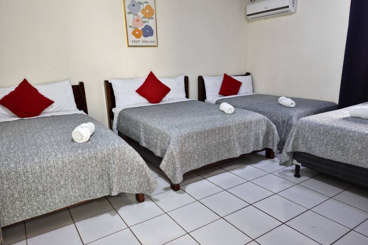Ein Hotelzimmer mit drei Betten, jedes mit grauer Bettdecke und roten Kissen. An der Wand hängt ein gerahmtes Kunstwerk und über den Betten befindet sich eine Klimaanlage.