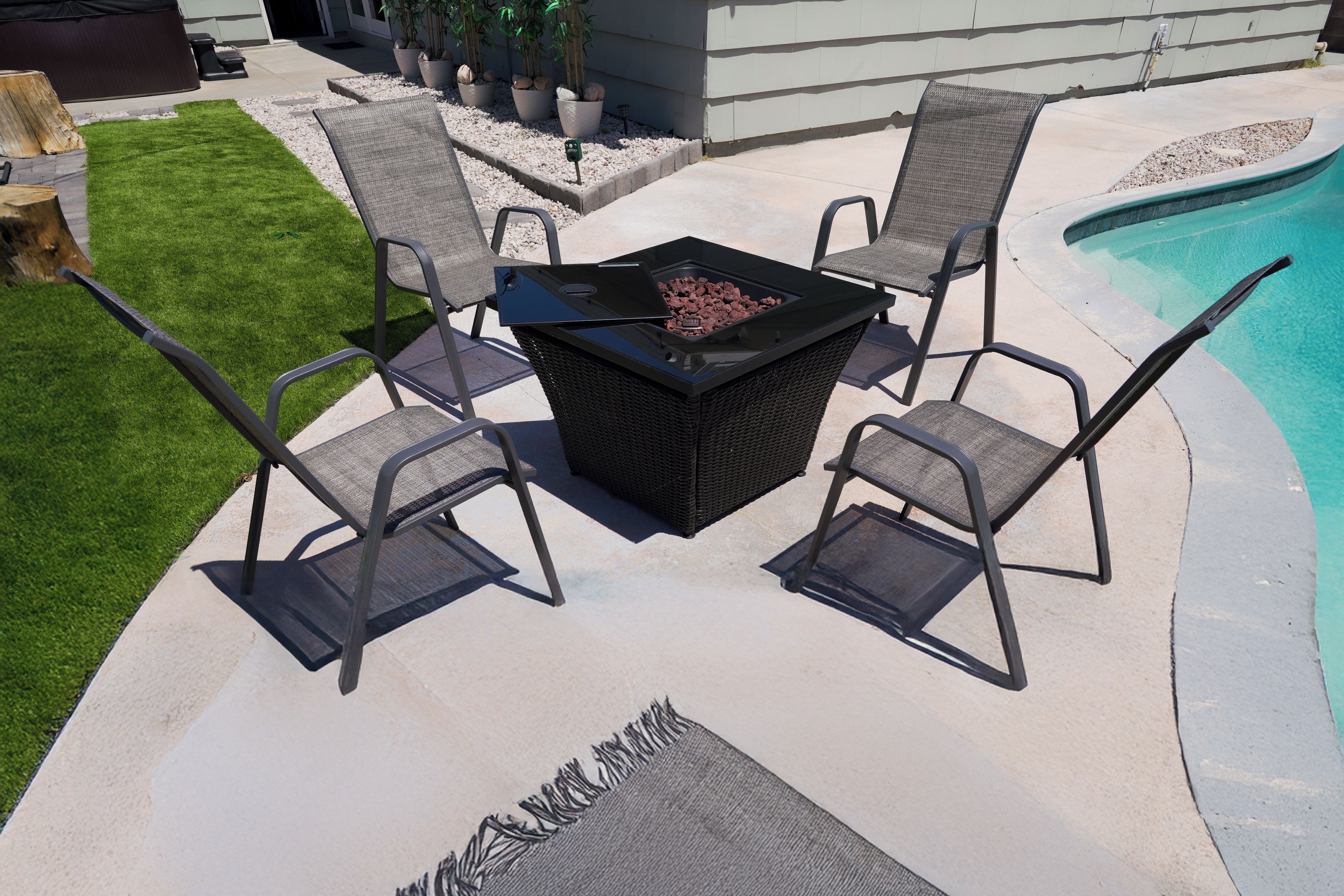 An outdoor patio with a black fire pit table surrounded by four chairs, a swimming pool, and a well-maintained lawn.