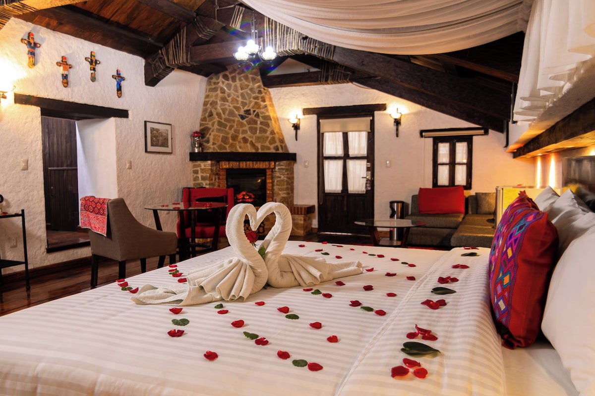 Una habitación acogedora con chimenea de piedra, una cama decorada con pétalos de rosa y un cisne hecho con toallas, y un comedor con una silla roja.