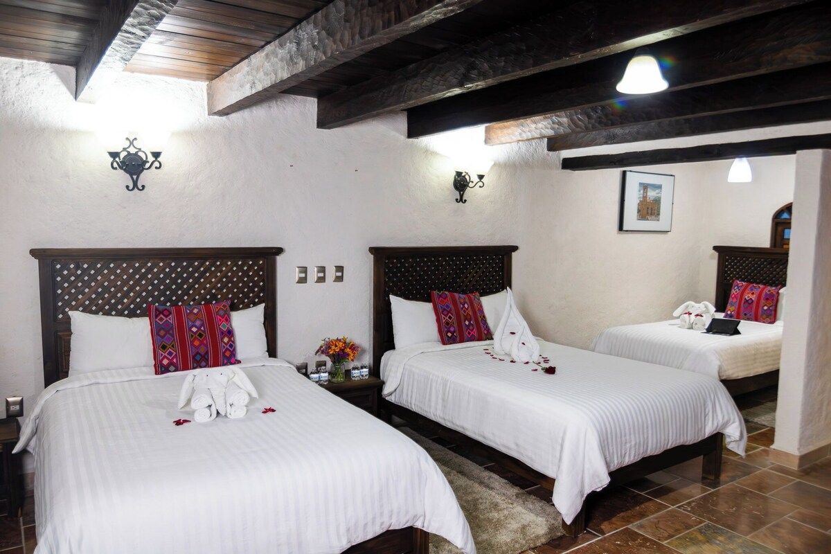 A hotel room with three beds, each with a wooden headboard and white bedding. There are wall-mounted light fixtures and a framed picture on the wall.