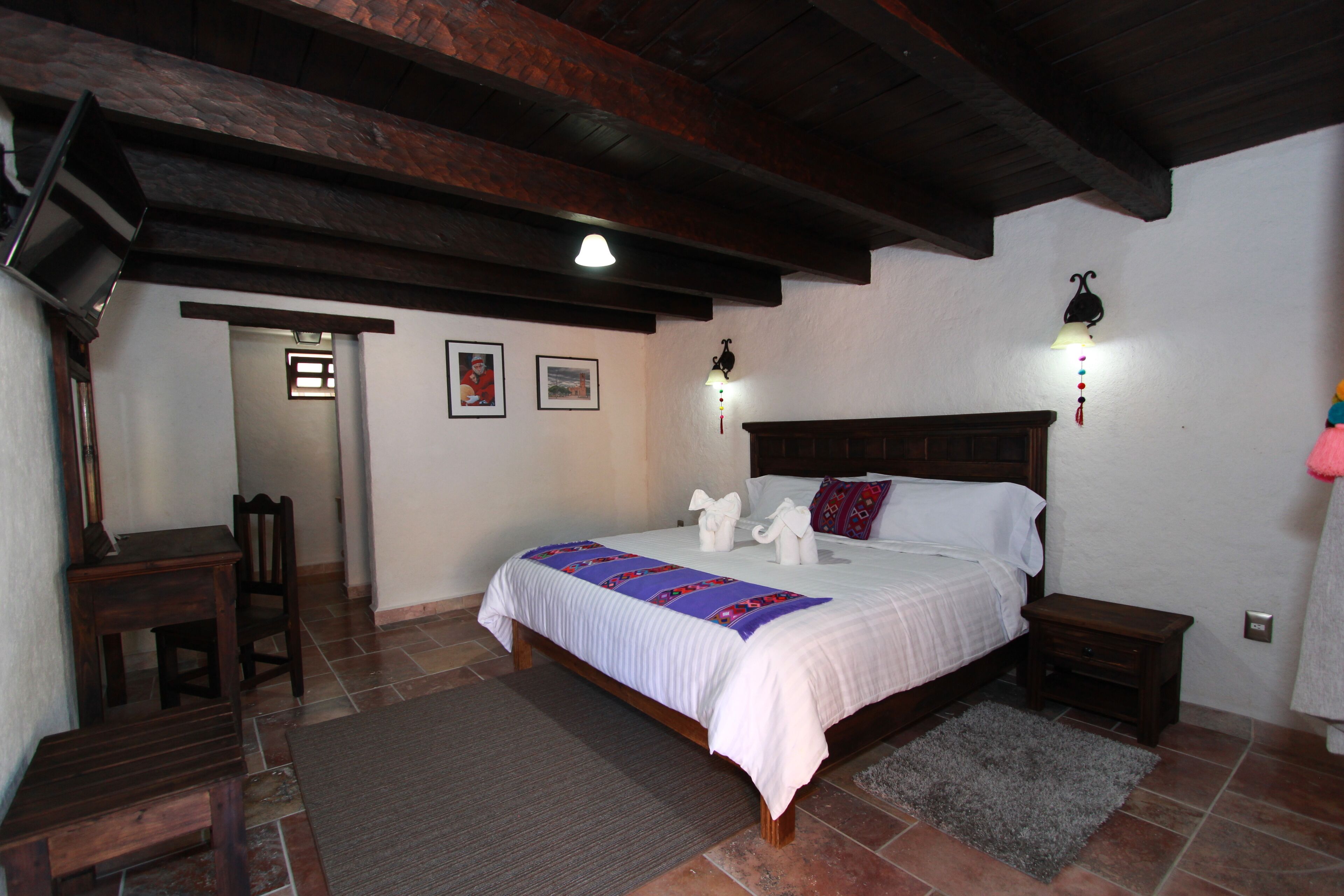 A bedroom with a wooden bed frame, a nightstand, a desk, and a chair. There are two wall-mounted lights and a ceiling light.