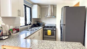 Una cocina con encimeras de granito, estufa a gas, campana extractora de acero inoxidable, refrigerador y ventana con vista a edificios.