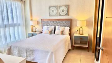 Habitación de hotel con una cama grande, dos mesitas de noche con lámparas, una ventana con cortinas traslúcidas y dos elementos decorativos en la pared.