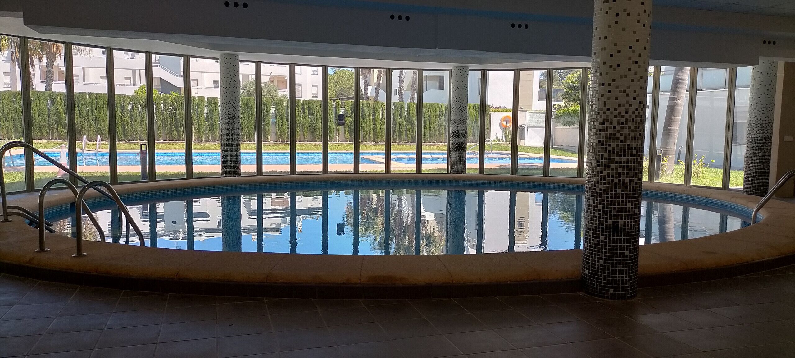 Ein Hallenbad mit gefliestem Boden, einer Metallleiter und Blick auf eine angelegte Grünfläche mit Bäumen und einem Gebäude im Hintergrund.
