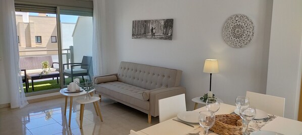 Una sala de estar moderna con un sofá color beige, una mesa de comedor con vasos y una vela, y un balcón con una silla y una mesita.