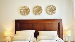 Un lit bien fait, avec du linge de lit blanc et deux oreillers, une tête de lit en bois et trois œuvres d’art murales en fibres tressées accrochées au-dessus.