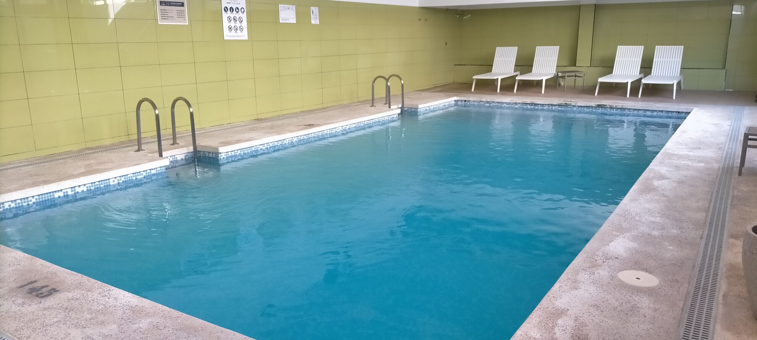 Una piscina interior rectangular con agua azul cristalina, rodeada por un muro de azulejos y algunas sillas reclinables blancas.