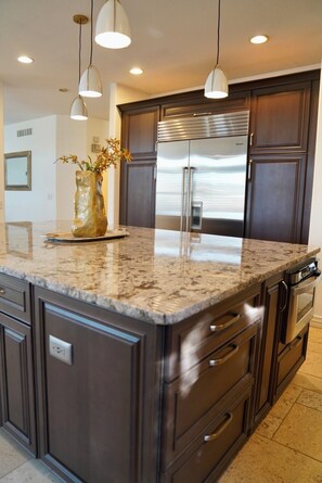 A kitchen with dark cabinetry, a stainless steel refrigerator, a granite countertop, and pendant lighting.