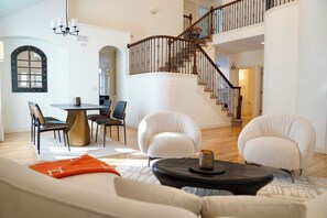 A spacious living room with a high ceiling, a staircase with a wooden railing, and a dining area with a round table and chairs.