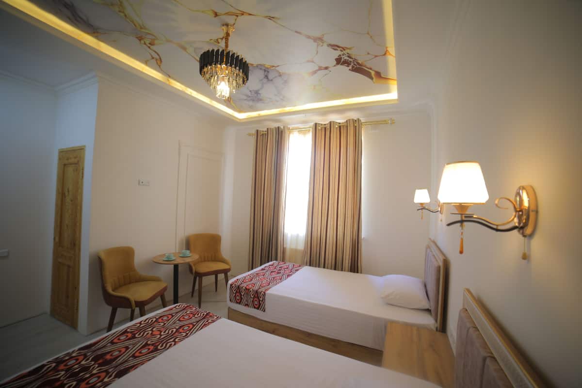 A hotel room with two beds, a small table, two chairs, a chandelier, and a wall-mounted lamp.