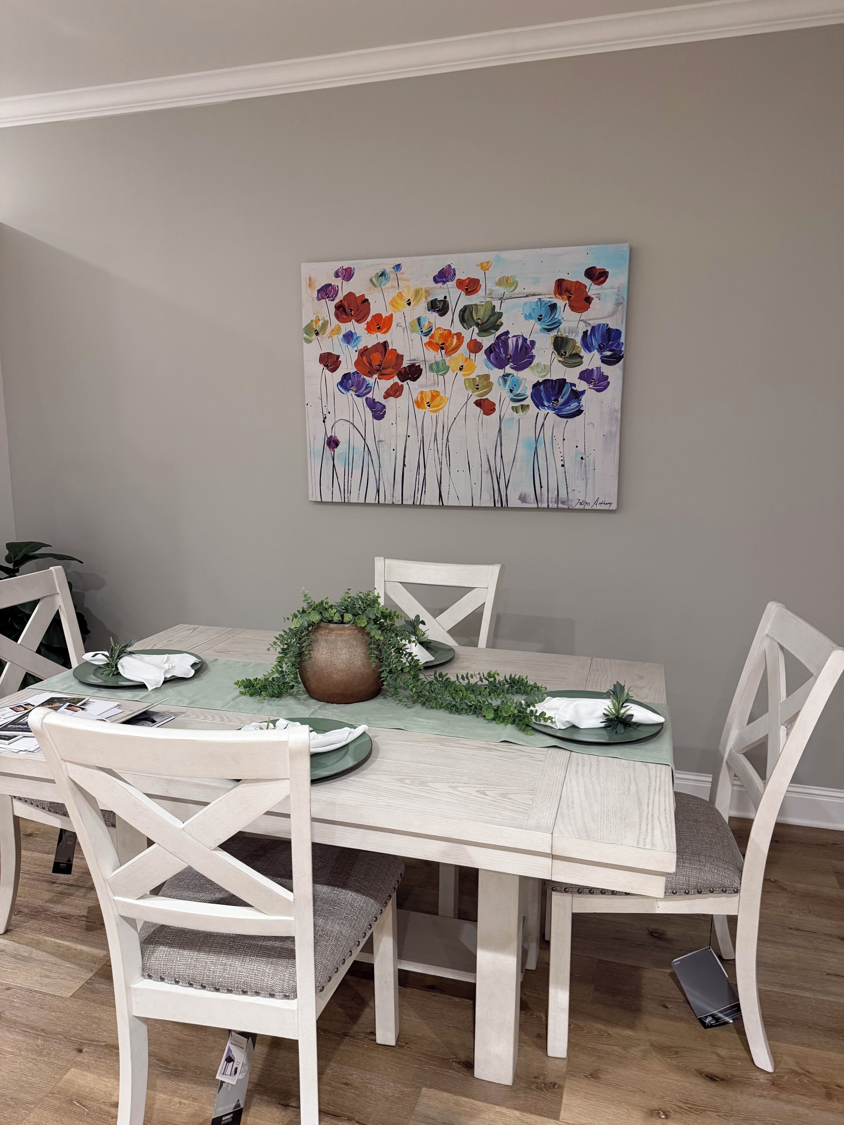 Un coin repas avec une table dressée pour quatre personnes, entourée de chaises blanches dotées d’assises rembourrées. Un tableau floral aux couleurs vives est accroché au mur, au-dessus de la table.