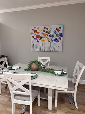 A dining area with a table set for four, surrounded by white chairs with cushioned seats. A colorful floral painting is on the wall above the table.