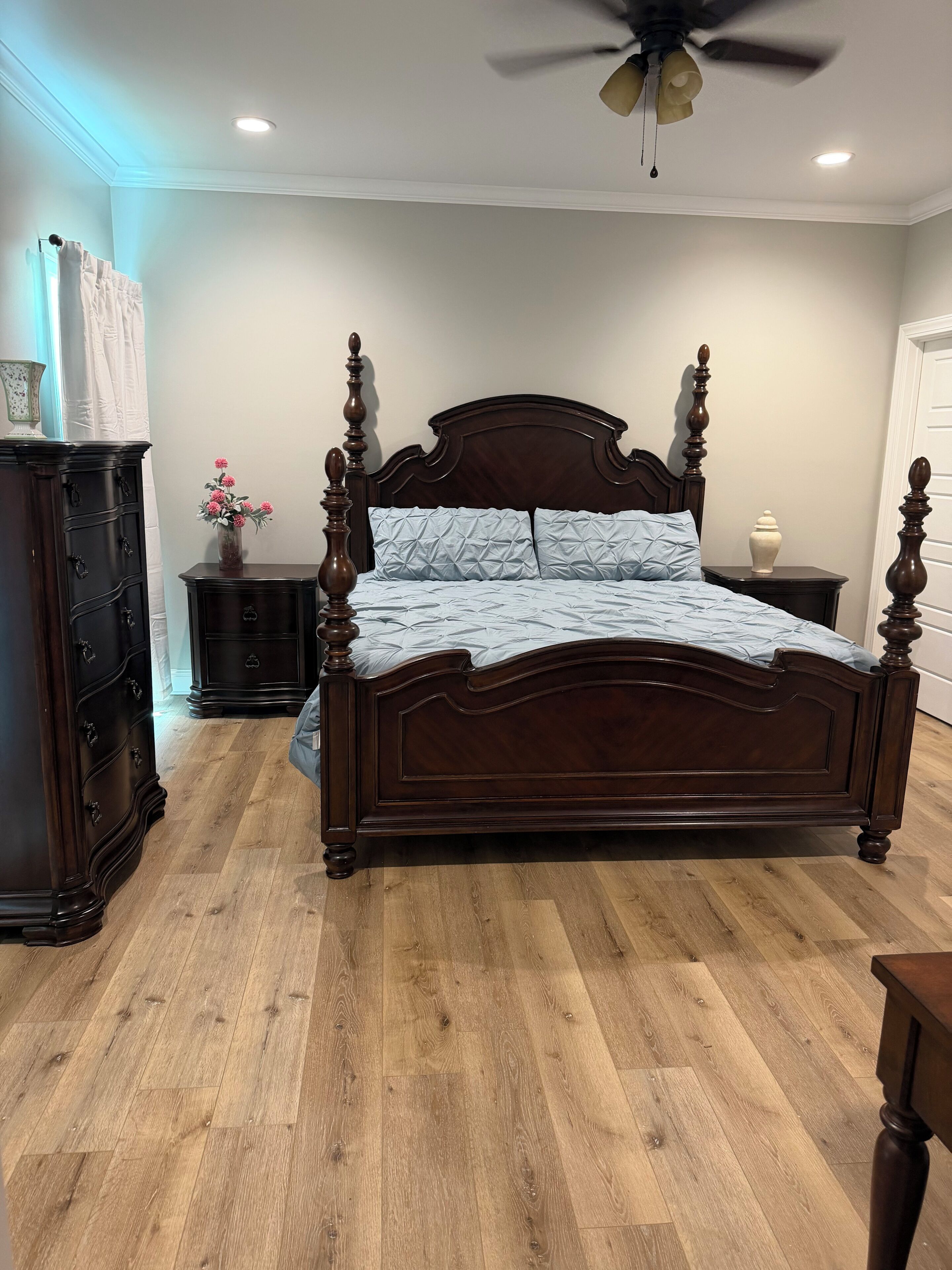 Een slaapkamer met een bed met houten baldakijn, een dressoir, een nachtkastje en een plafondventilator.