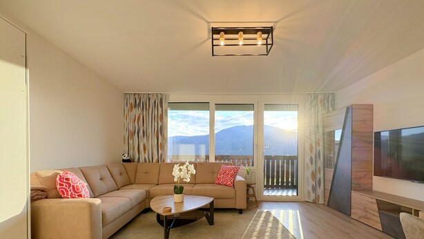 Una sala de estar moderna con un sofá color beige, una mesa de centro y un televisor. Se ven ventanas amplias con vistas al exterior.
