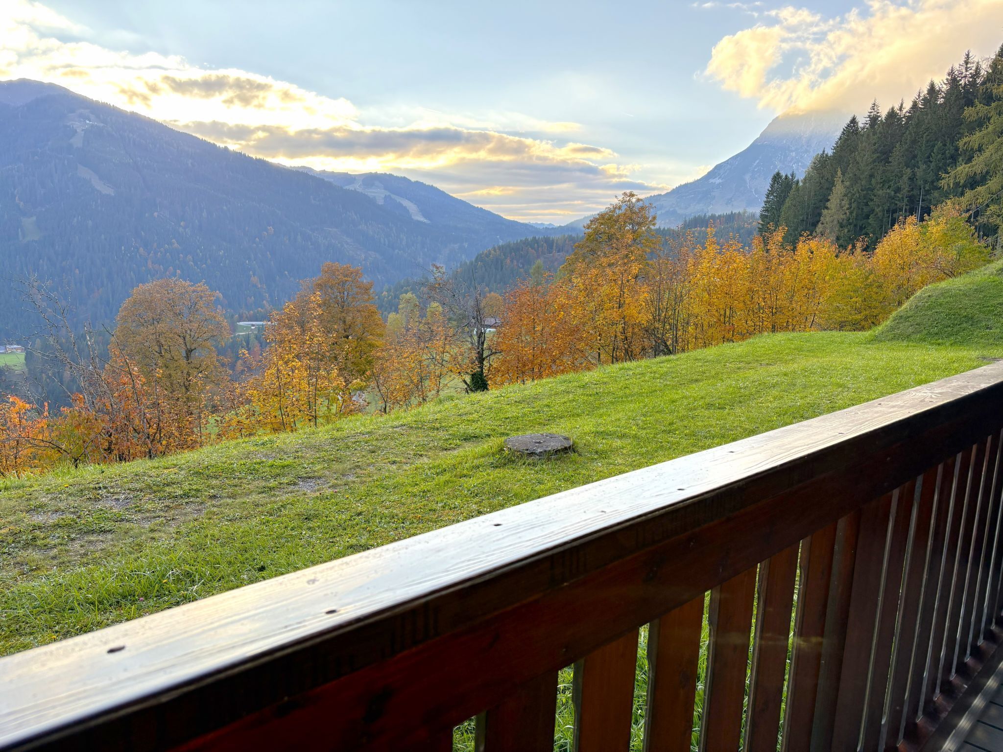 A scenic view from a wooden deck overlooking a grassy hillside with autumn-colored trees and mountains in the background.