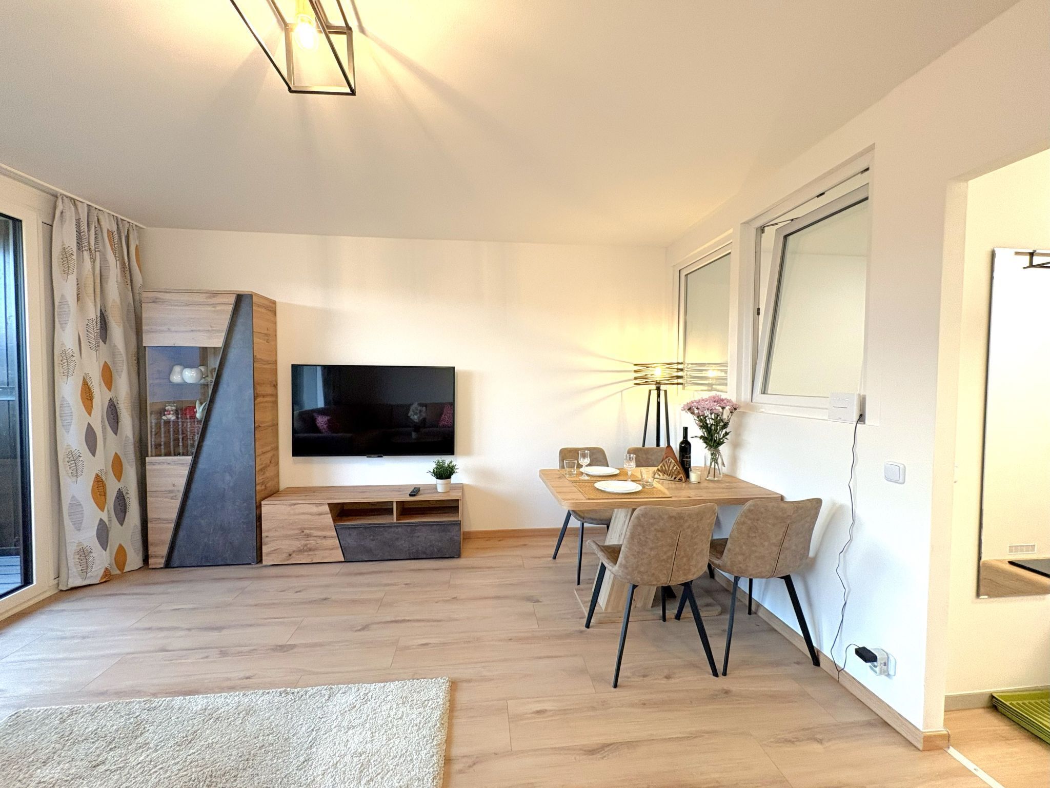 Un salon moderne doté d’un téléviseur à écran plat, d’un coin repas avec une table dressée pour deux personnes et d’une grande fenêtre agrémentée de rideaux.