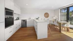 A modern kitchen with white cabinetry, a built-in microwave, and a dishwasher. There is a dining area with a wooden table and white chairs. - Spacious 4BR w/ Water Views (Port Macquarie)