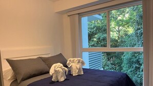 A neatly made bed with a dark blue bedspread and two pillows, a window with blinds, and a view of greenery outside. - Great Apartment Punta Esmeralda (Puntarenas Province)