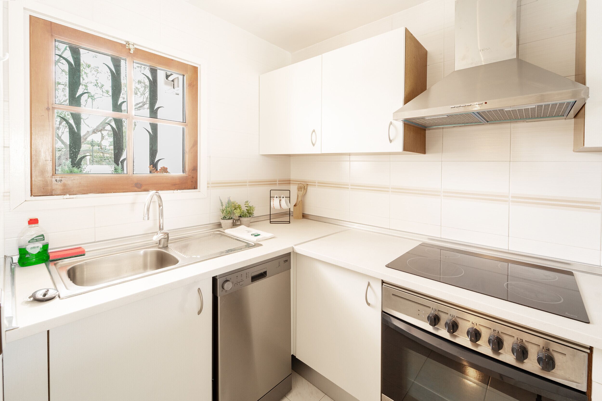 A modern kitchen with white cabinets, a dishwasher, a stove, and a window with a view of trees.