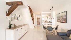 A modern living room with a gray sofa, a dining area with a white table and chairs, a kitchen in the background, and a staircase with wooden beams. - WELCS CASA ADOSADA 215 EMP con piscina comunitaria (Rosas / Roses)