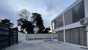 A rooftop parking area with a concrete surface, a metal fence on one side, and a building with large windows and an air conditioning unit on the other.