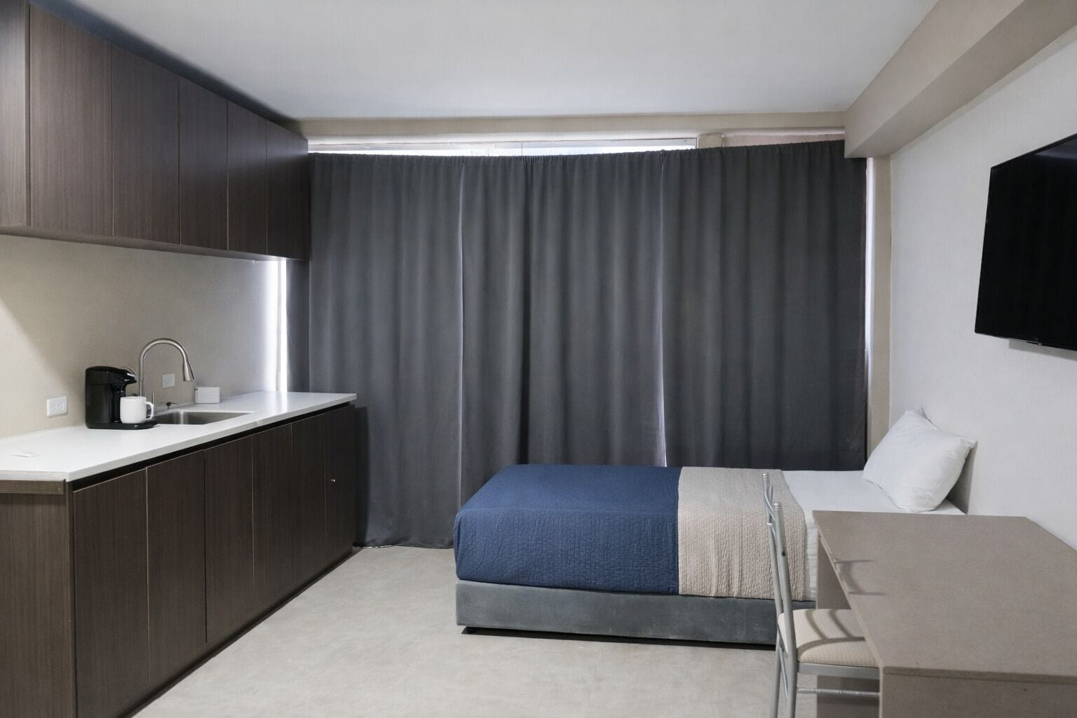 Habitación de hotel pequeña con kitchenette, cama con manta azul, televisión montada en la pared y ventana con cortinas.