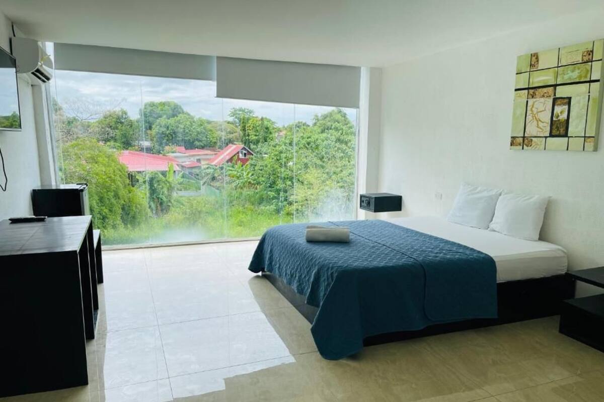 A modern bedroom with a large window, a bed with a blue bedspread, a desk, and a wall-mounted artwork.