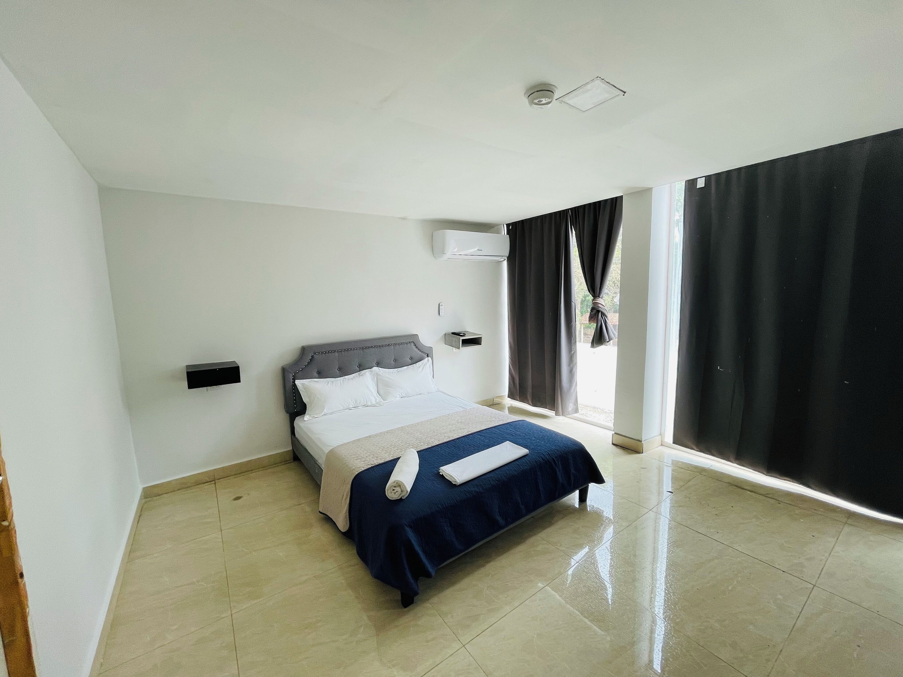 A hotel room with a bed, a window with curtains, a ceiling light, and a wall-mounted air conditioning unit.