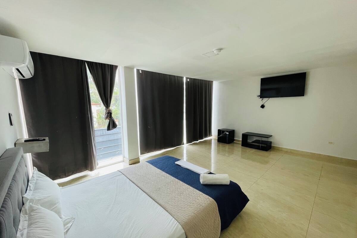 A hotel room with a bed, a flat-screen TV, a ceiling fan, and a window with curtains.
