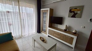 Un salon moderne avec un canapé, une table basse, un téléviseur posé sur un meuble en bois et une grande fenêtre ornée de rideaux légers.