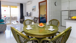Un coin repas avec une table ronde et quatre chaises, une cuisine équipée d’un réfrigérateur et d’une horloge, et un salon avec un canapé et un téléviseur.
