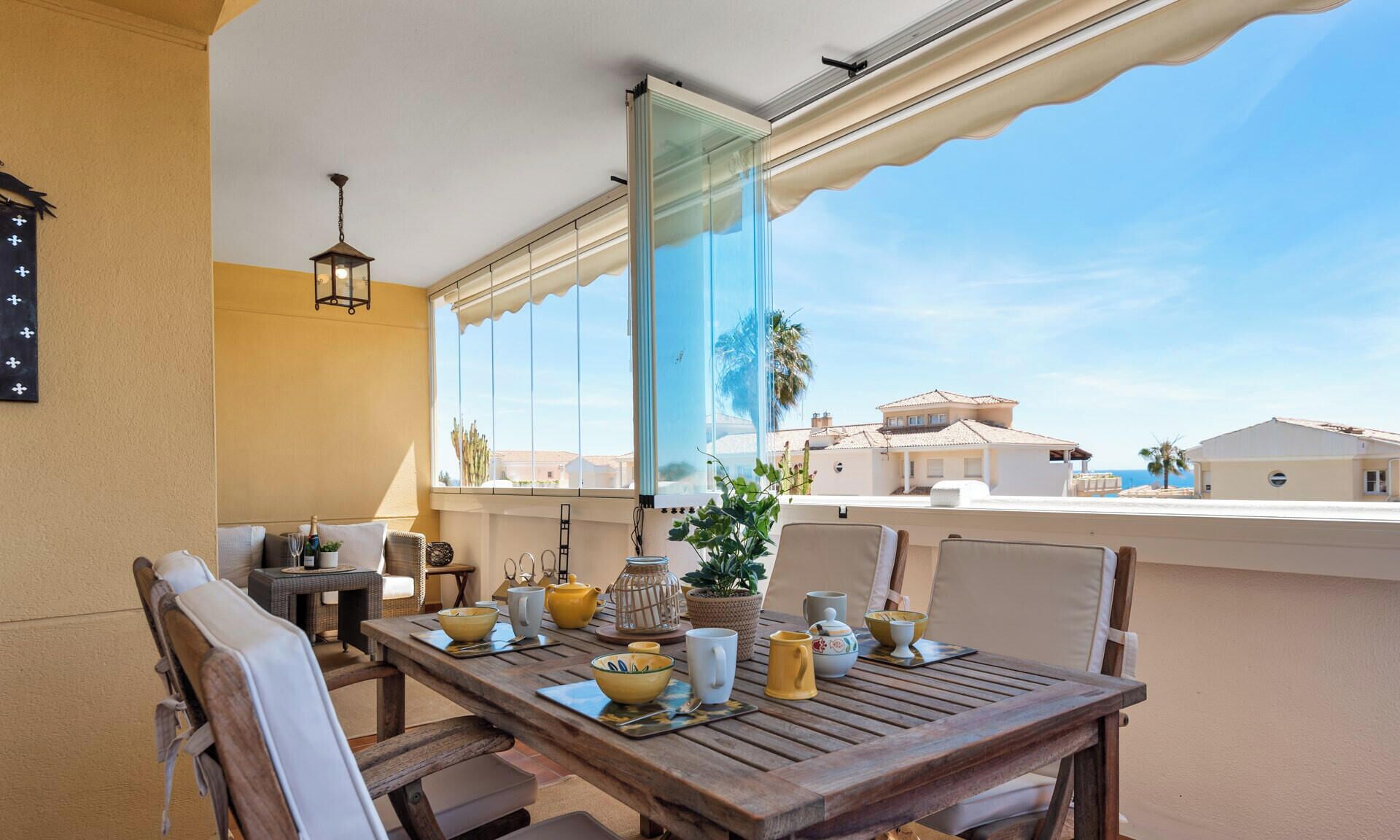 Een houten eettafel met stoelen, een plant in pot en divers keukengerei op een balkon met uitzicht op gebouwen en palmbomen.