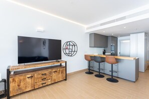 A modern living space with a flat-screen TV, wooden entertainment unit, kitchen area, and bar stools.