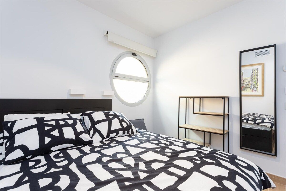 A bedroom with a bed, a round window, a wall-mounted shelf, and a framed picture.