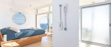 A modern bedroom with a bed, a round window, and a view of the sea.