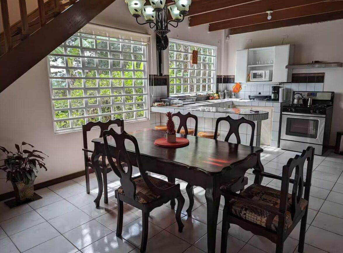 Uma área de refeições com mesa e cadeiras de madeira, cozinha ao fundo e grandes janelas com vista para o verde.