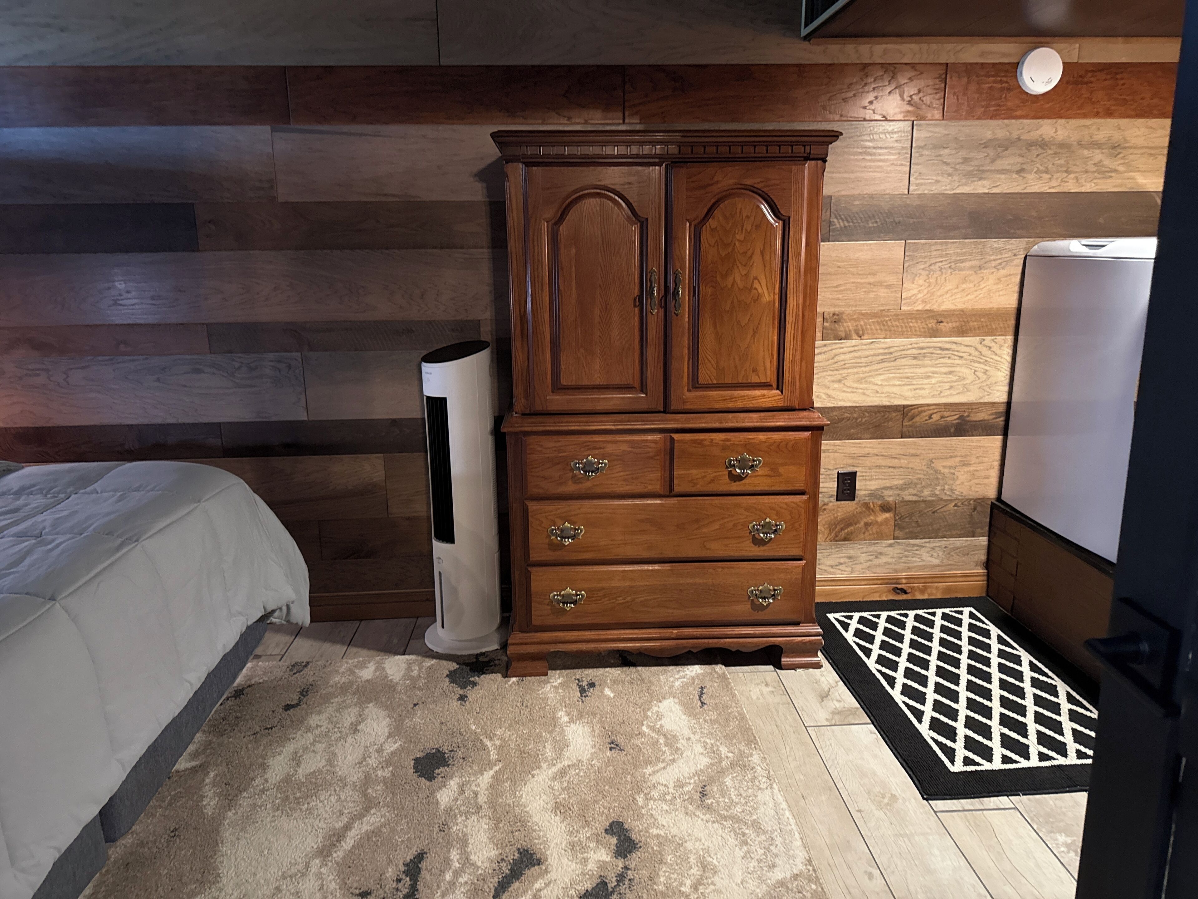 A bedroom with a wooden wardrobe, a standing heater, a bed with a white duvet, and a patterned rug.