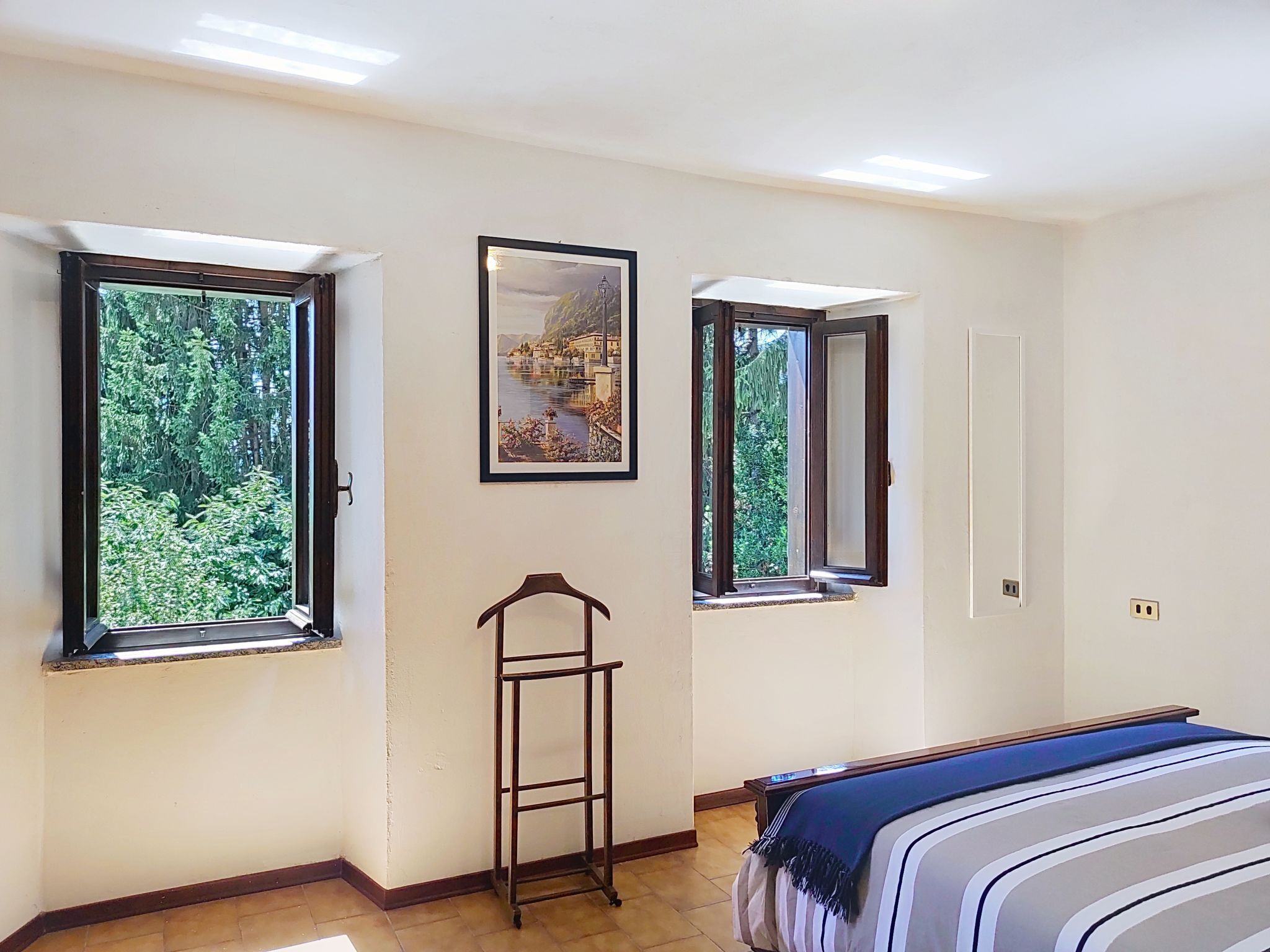 A bedroom with a bed, two windows with views of greenery, a framed picture, and a small wooden ladder.