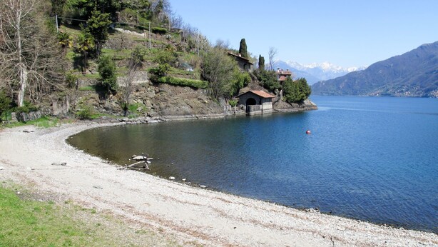 Une vue sur un lac, avec un petit bâtiment situé sur une rive rocheuse, entouré d’arbres et avec des montagnes en arrière-plan.