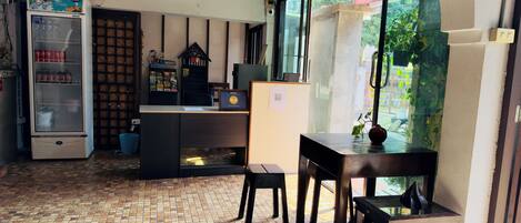 Un café acogedor con suelo de baldosas, un refrigerador lleno de bebidas, una barra de madera y vistas al exterior con vegetación.