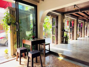 An open-air seating area with a table and two stools, a glass door leading outside, and a concrete floor. - Mi Casa Thaphae Chiang Mai (Chiang Mai)