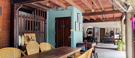 A rustic wooden interior with a long table and wicker chairs, a blue door, and a fan on the ceiling.