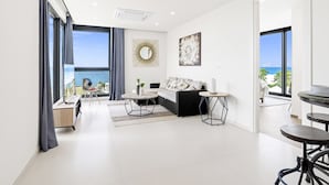 A modern living room with a sofa, coffee table, and dining table, large windows with a view of the sea, and a round mirror on the wall. - A601 (Cupecoy)